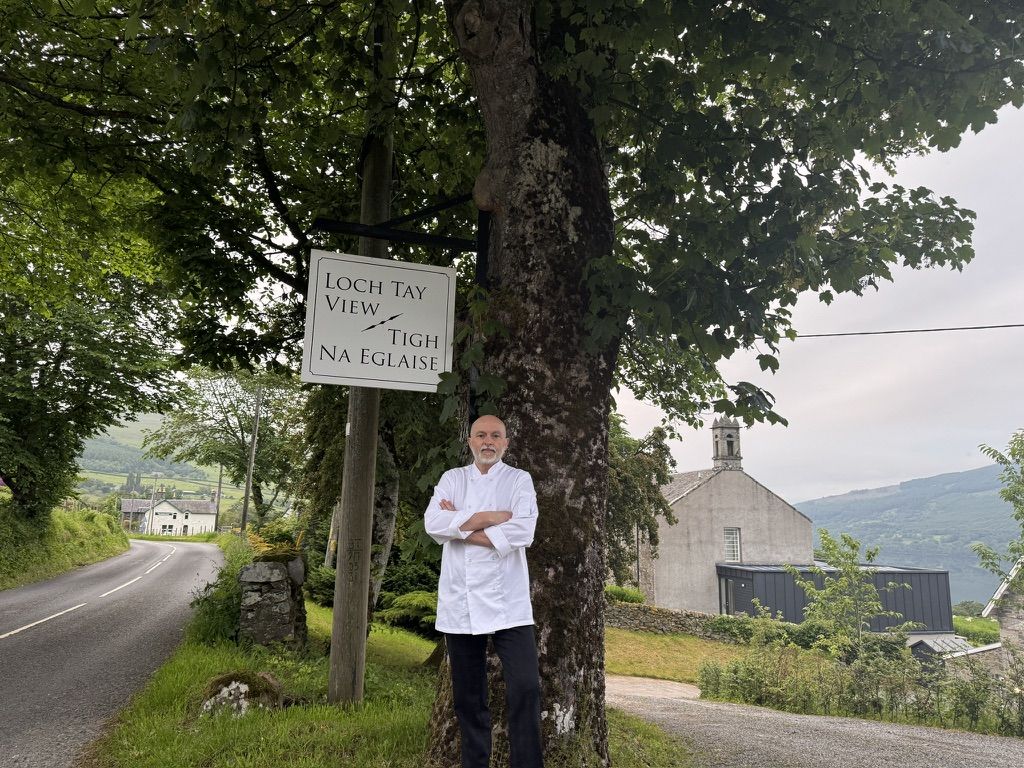 Private chef Giuseppe Manzoli by the Loch Tay View / Tigh na Eglaise sign, with the church and Perthshire hills behind.